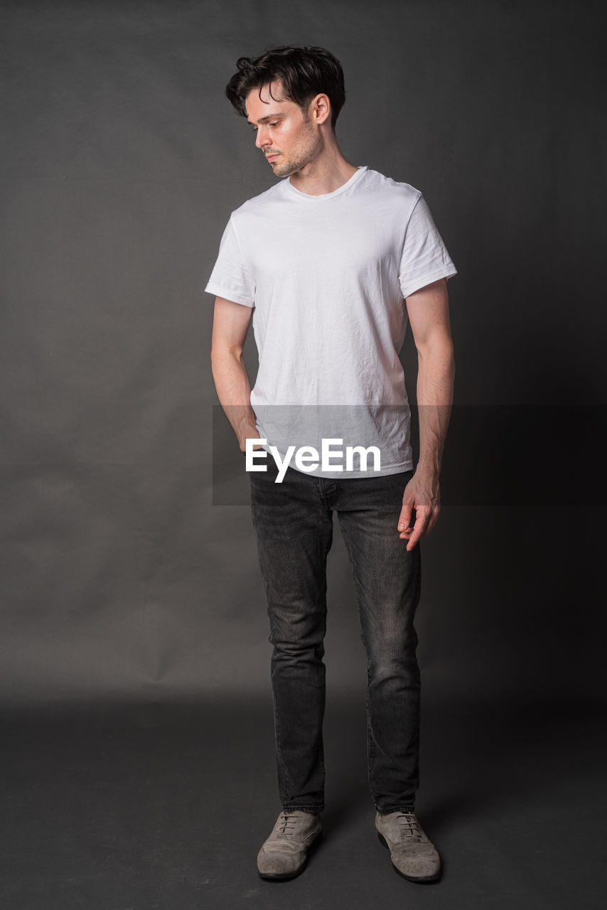 one person, t-shirt, studio shot, standing, casual clothing, young adult, adult, portrait, men, indoors, gray, photo shoot, full length, pocket, white, black, clothing, sleeve, gray background, front view, looking, fashion, looking at camera, emotion, hands in pockets, beard, facial hair, spring, smiling, cool attitude, copy space, brown hair, relaxation, individuality, lifestyles, trousers, jeans, outerwear