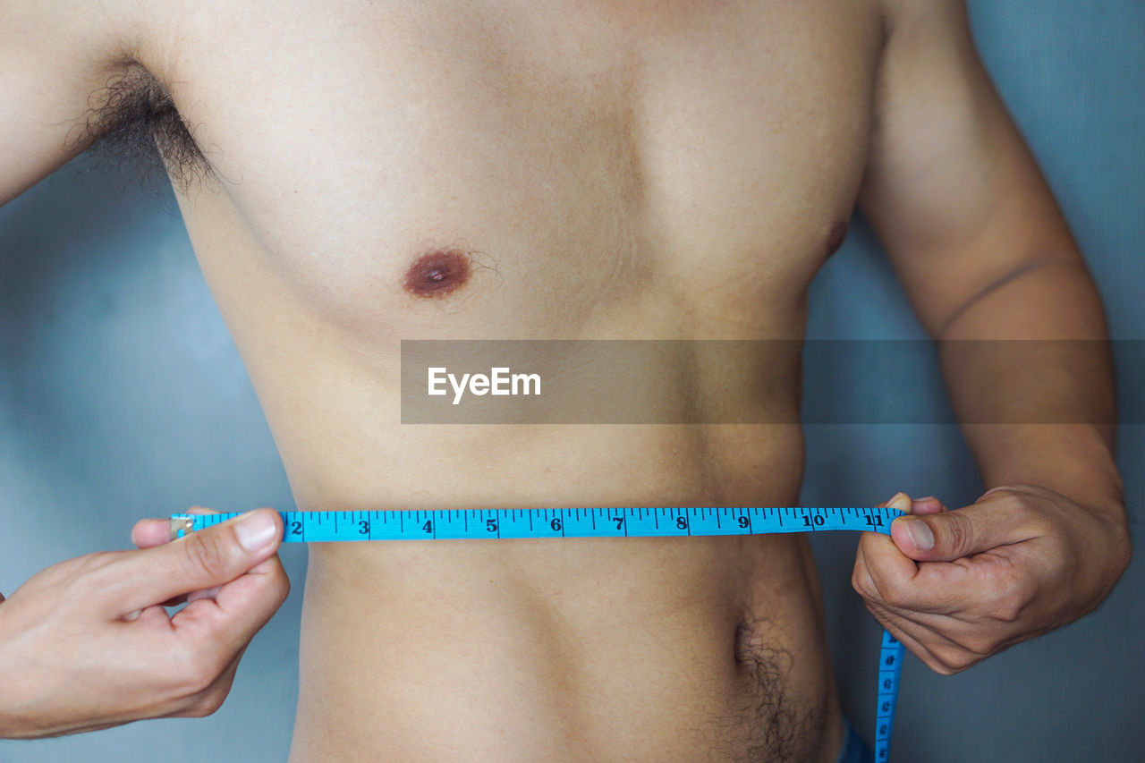 Midsection of shirtless man measuring waist with tape measure