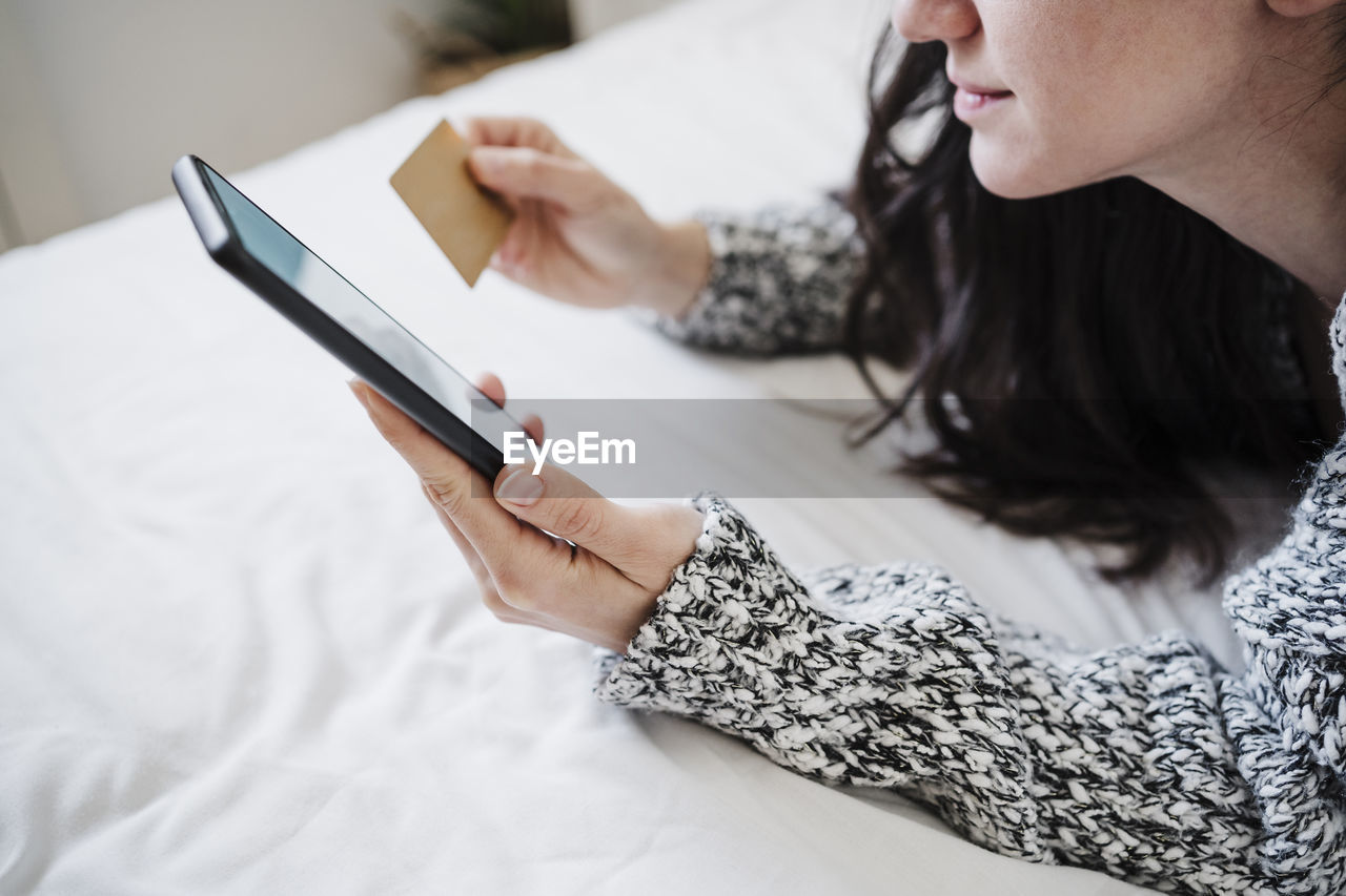 Woman with credit card doing online shopping through mobile phone while lying on bed at home