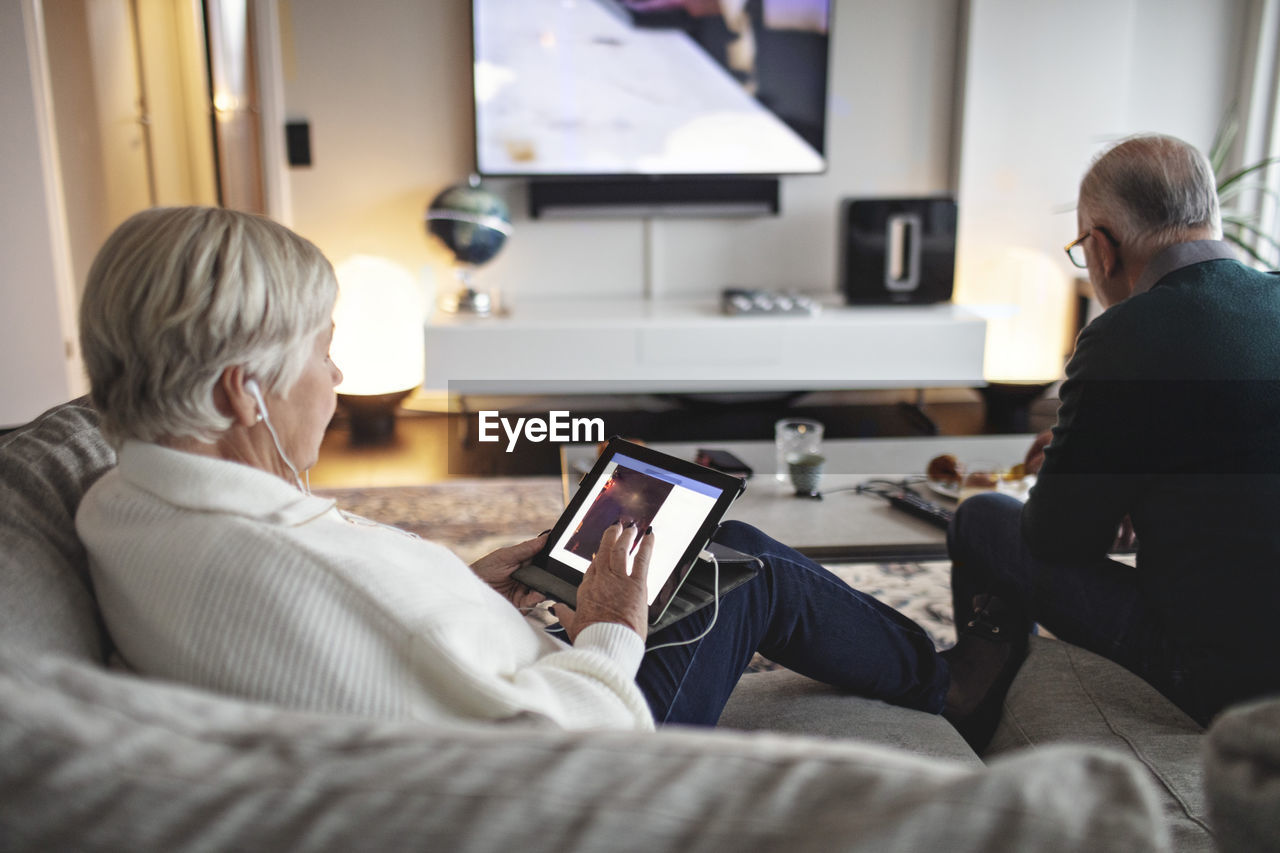 Senior woman using digital tablet while male partner sitting on sofa in living room