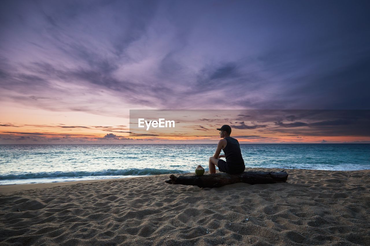 Side view of young man sitting on log at beach against sky during sunset