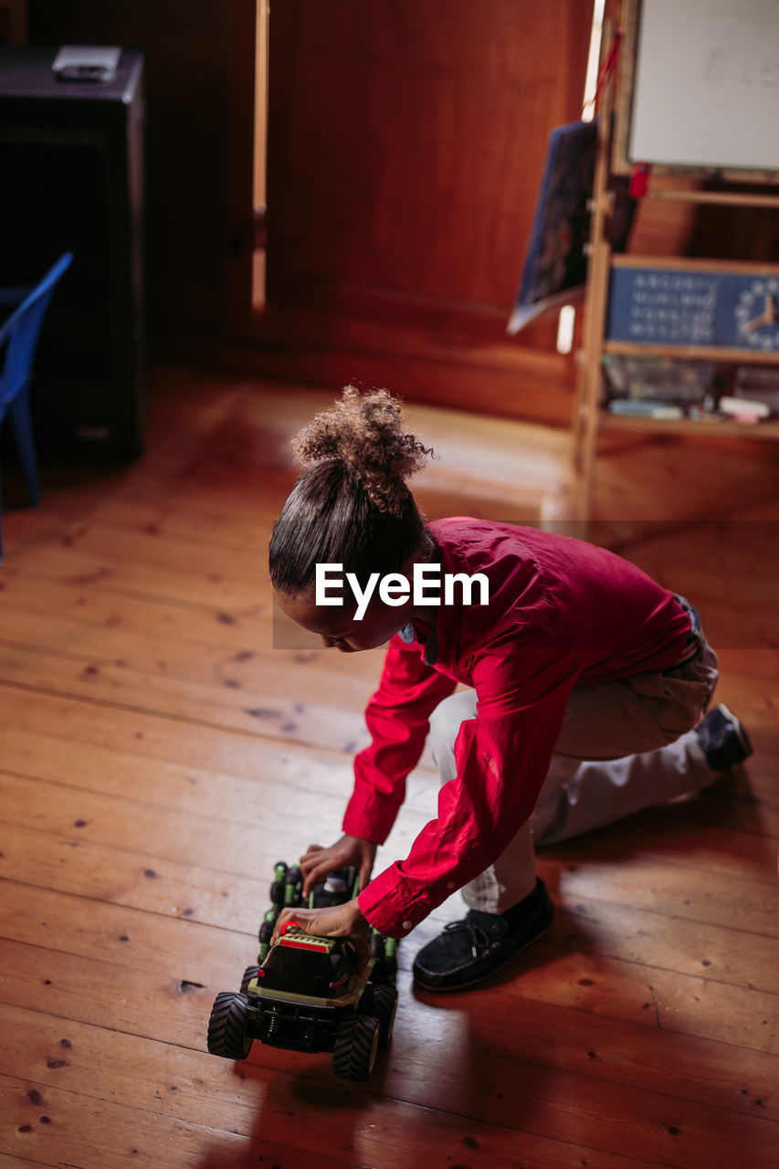 From above side view of ethnic child in casual clothes sitting on wooden floor and playing with toy car at home