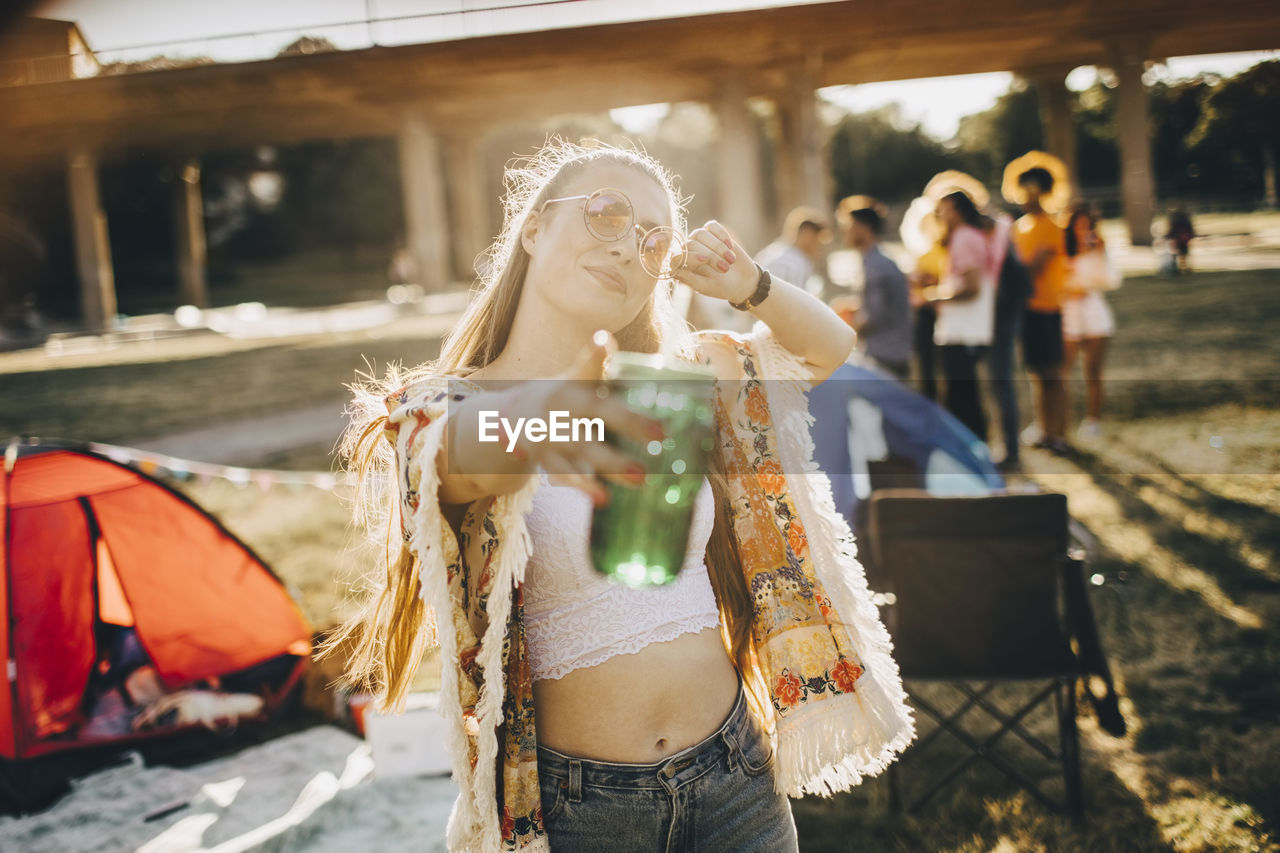 Portrait of playful young woman holding drink while pointing at event on sunny day