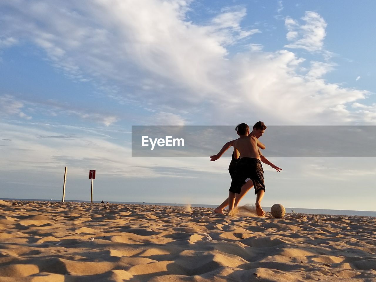 Full length of teenage boys playing soccer at beach against sky