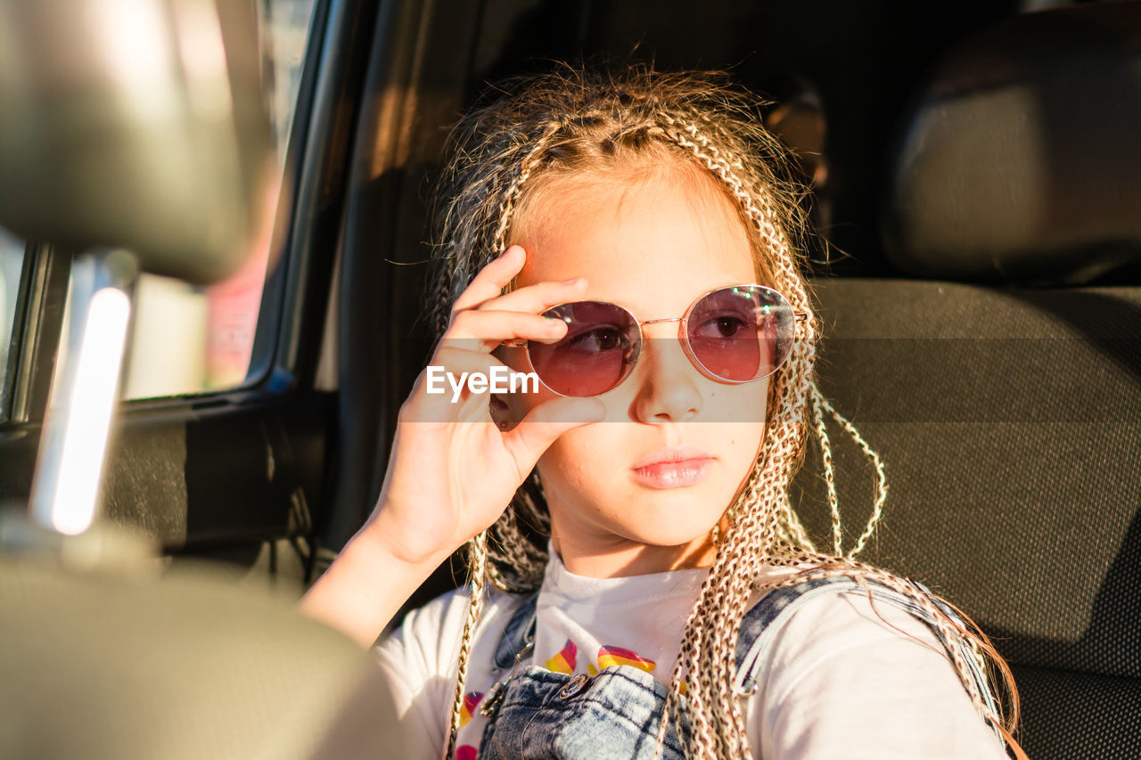 Serious teenager girl with afro-braids holds sunglasses with her hand and looks out the window 