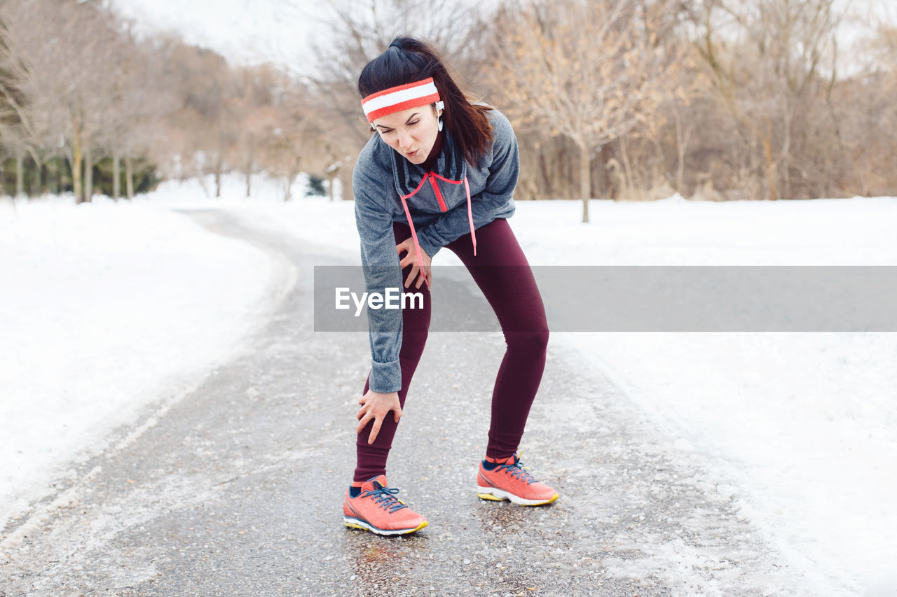 Full length of woman wearing sports clothing suffering from knee pain on road during winter