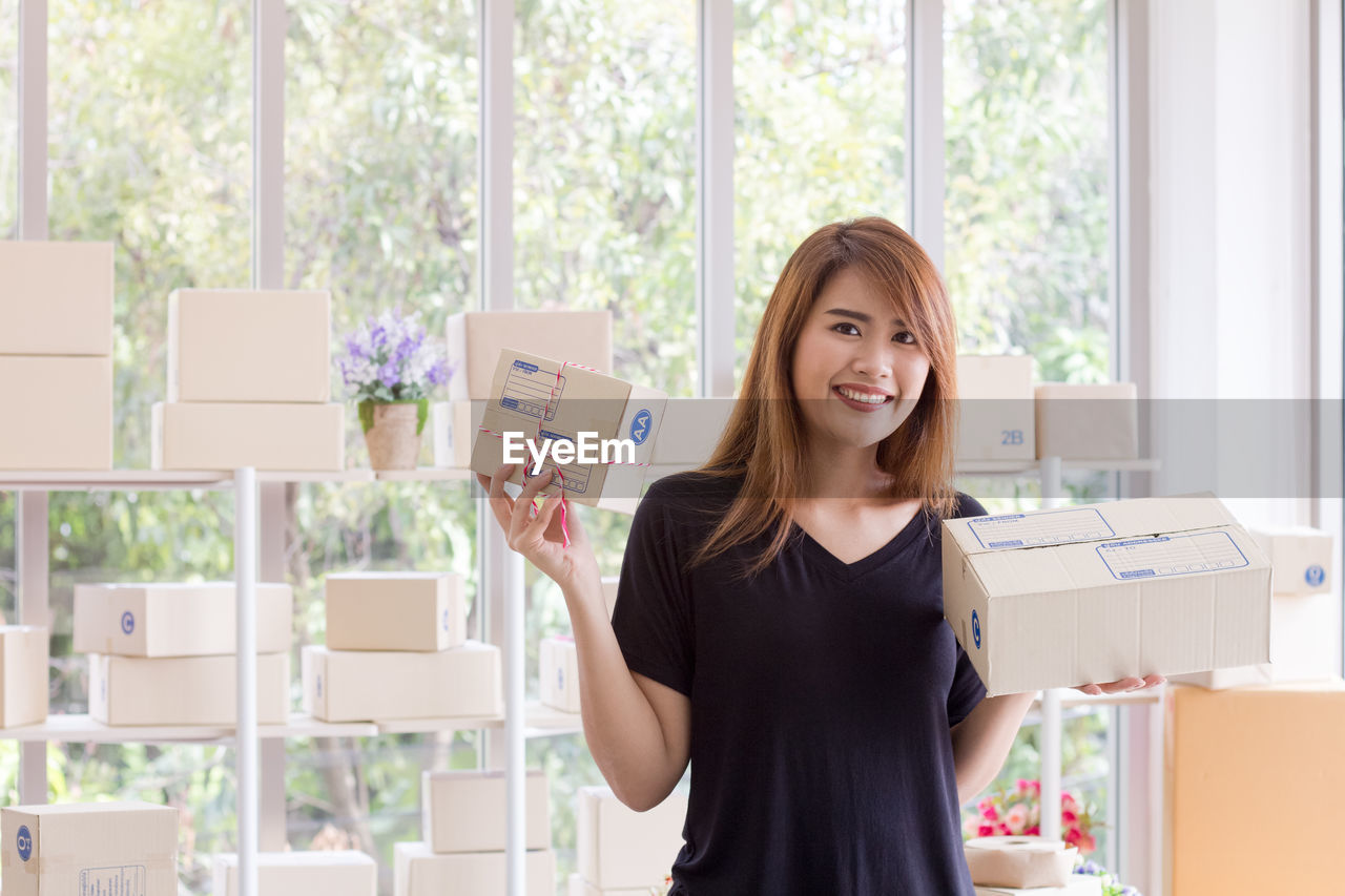 Portrait of smiling young woman holding cardboard boxes against window in office