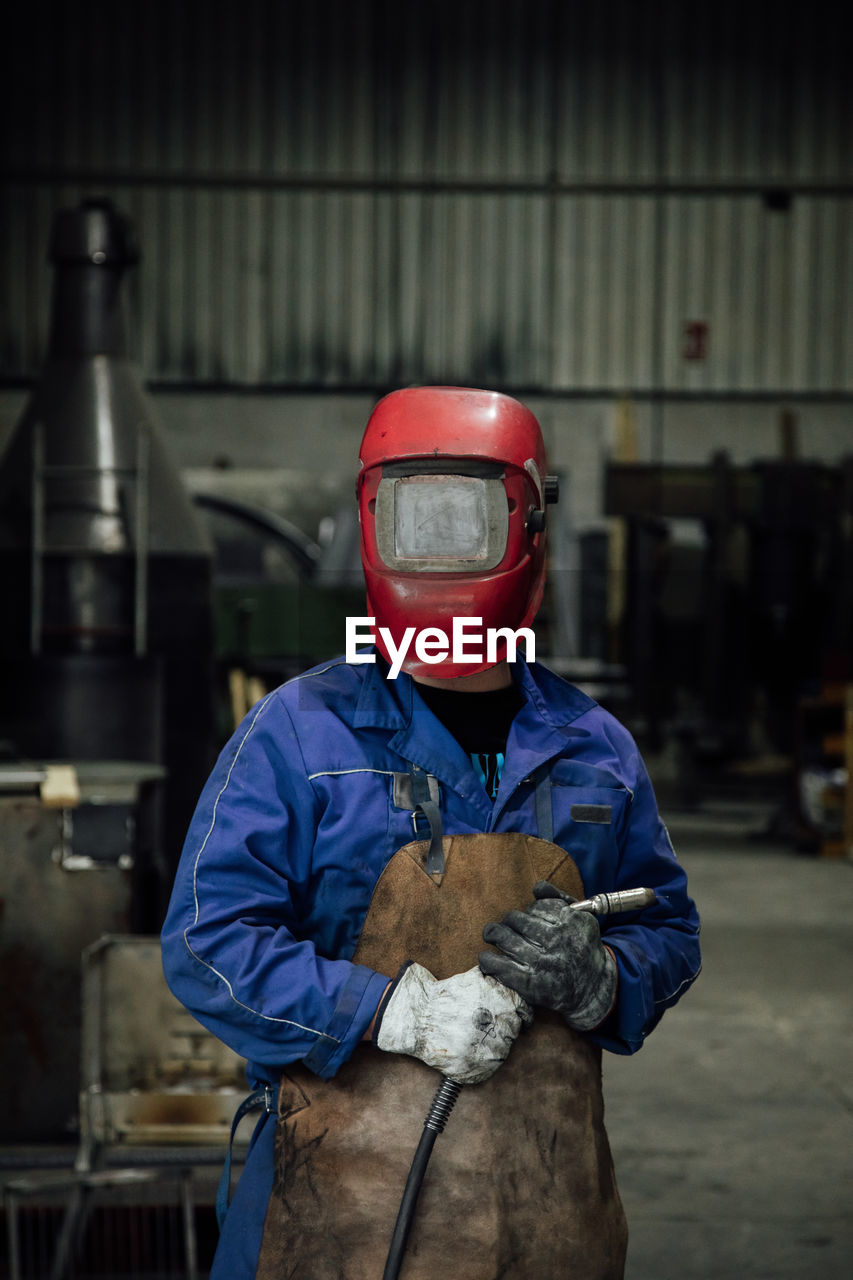 Anonymous mechanic in protective helmet and apron on blue overall standing with welding hose in light workshop near metal constructions person