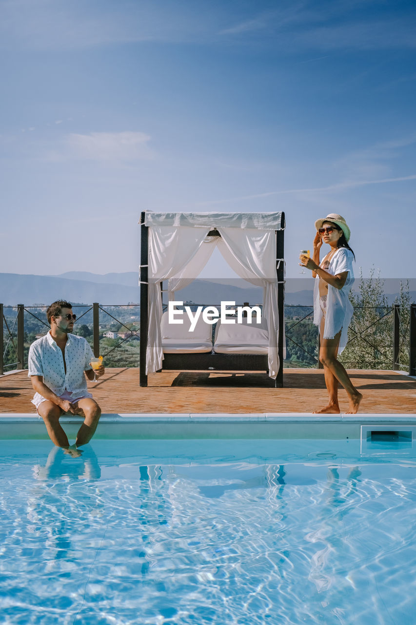 Couple sitting by swimming pool against sky