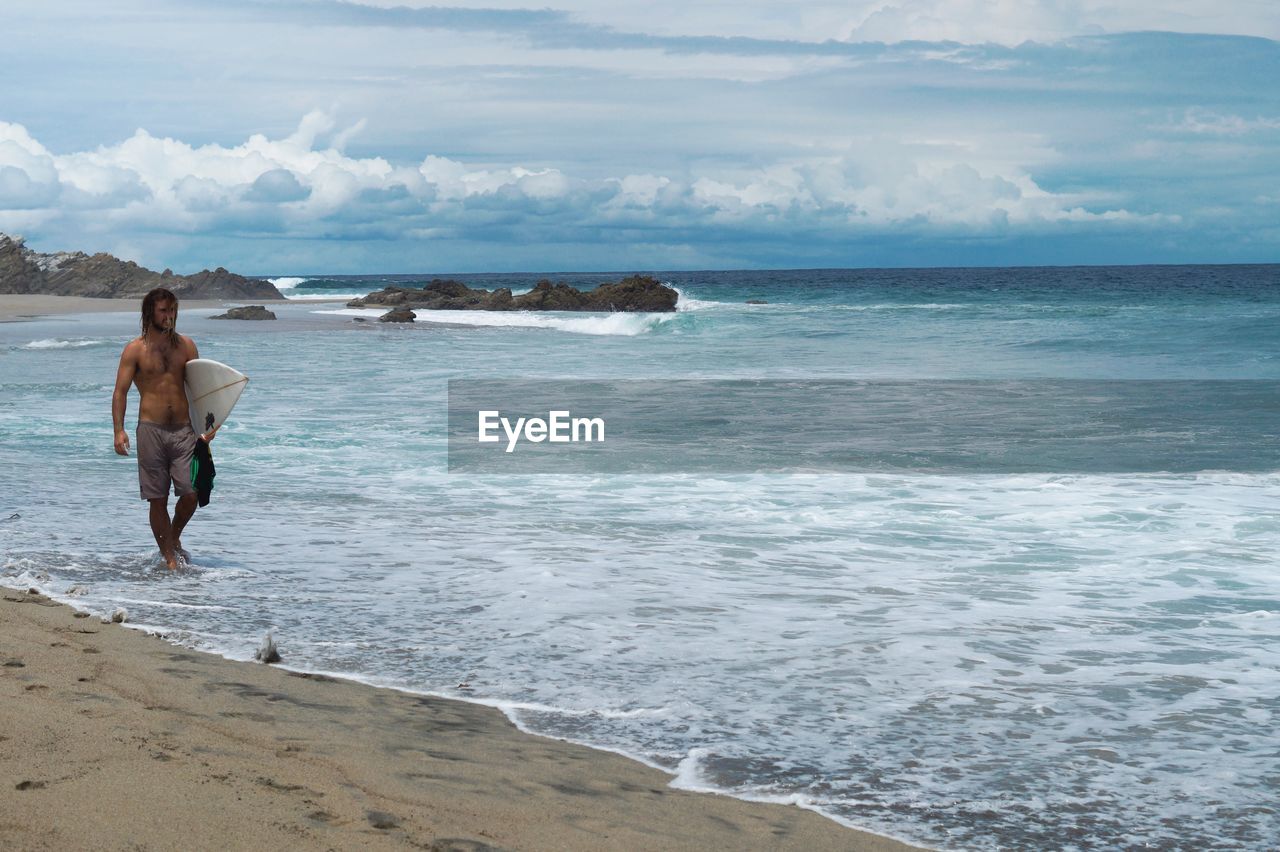 Full length of shirtless man carrying surfboard walking at shore against sky