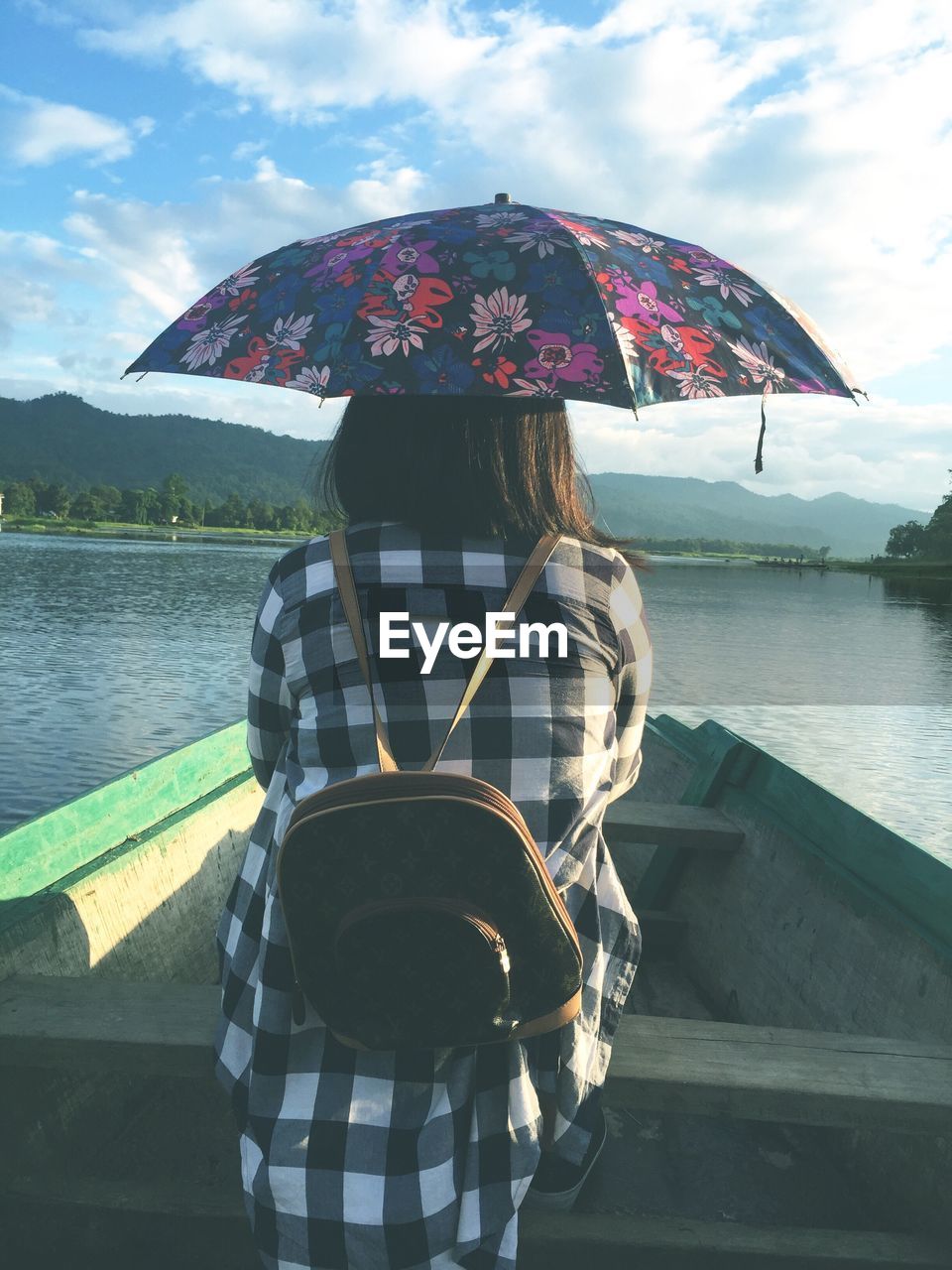 Rear view of woman holding umbrella while sitting in boat on lake against sky