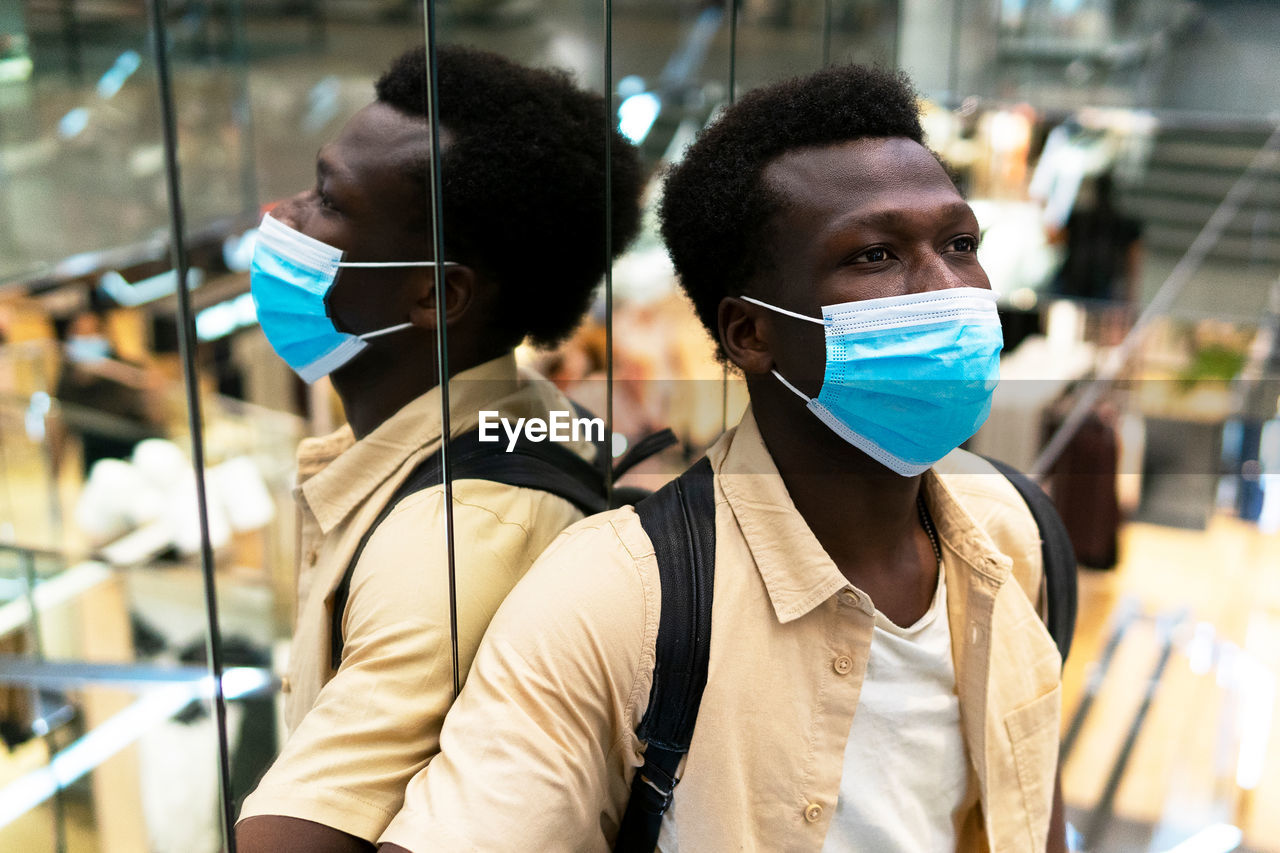 Calm african american male customer in medical mask standing near mirrored wall in modern shopping center and looking away during coronavirus