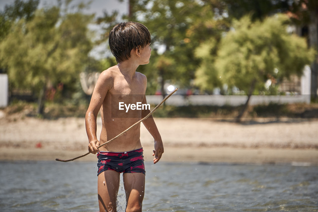 Shirtless boy holding stick while standing in lake