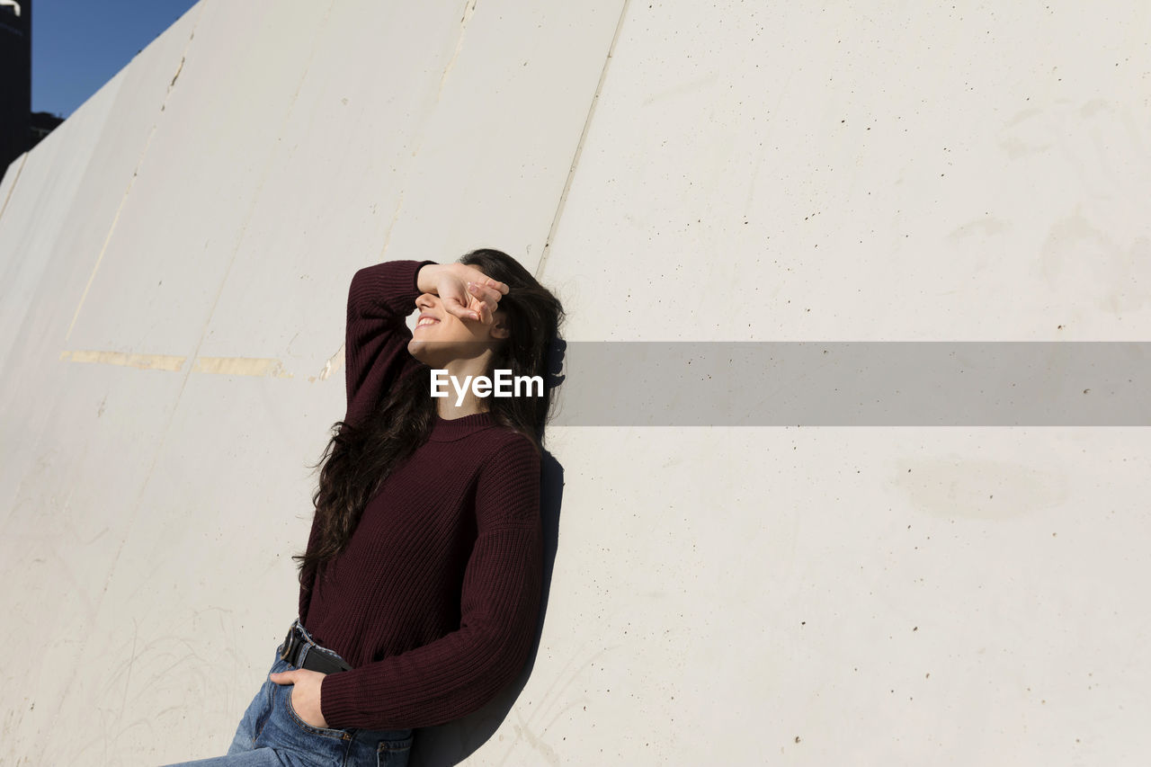 Woman with hands in pockets shielding eyes while leaning on wall during sunny day