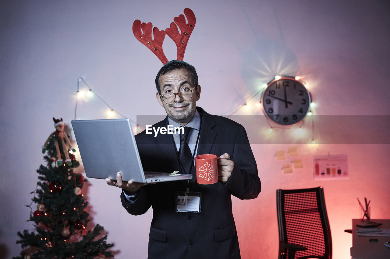 Portrait of businessman wearing headband holding laptop and coffee cup against illuminated christmas tree and lights at home