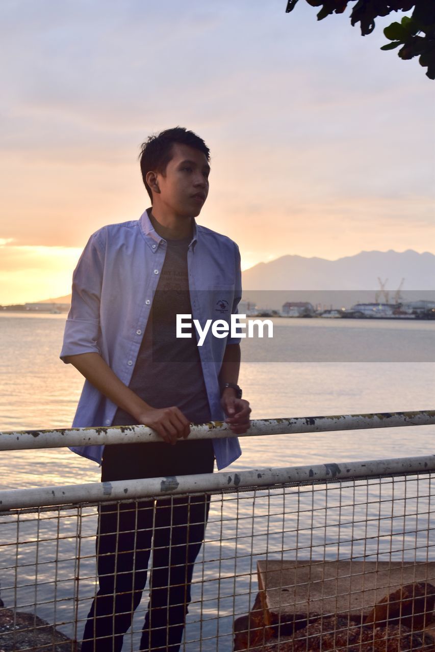 Man standing by railing against sea during sunset