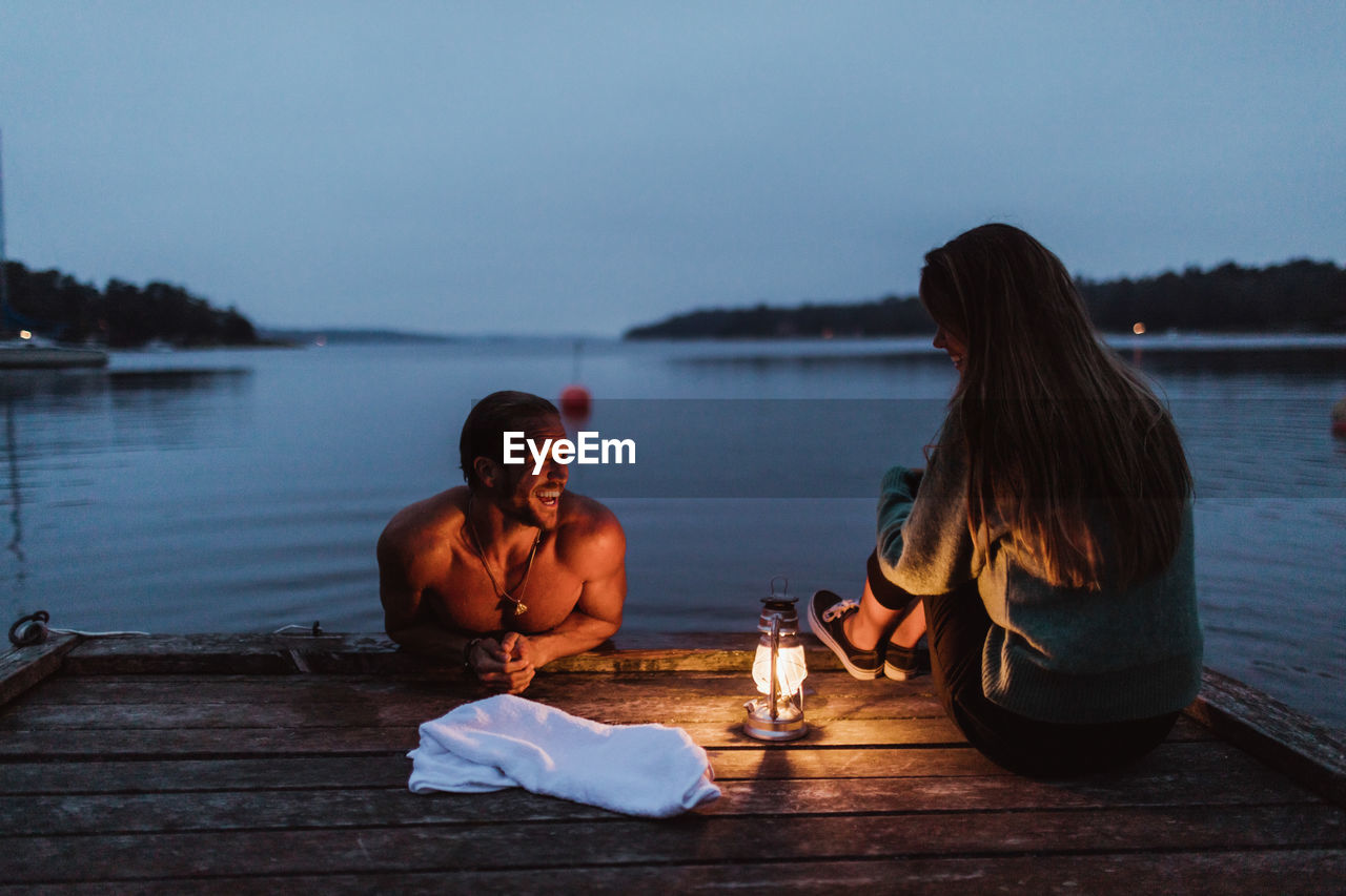 Smiling man in water talking with woman sitting on pier by sea against sky