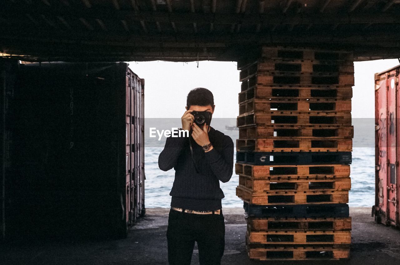 Man photographing through camera while standing against stacked crate