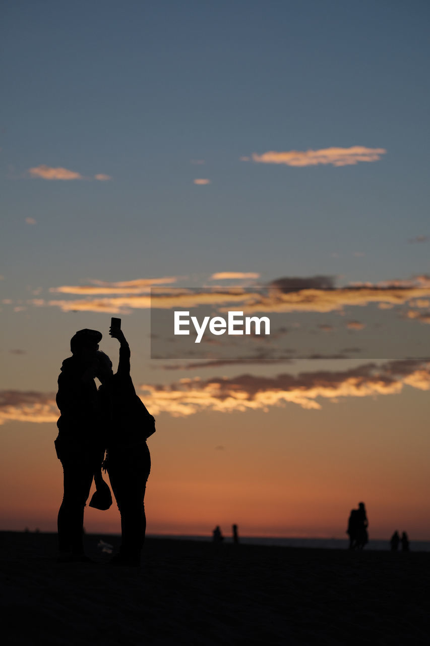 Silhouette couple taking selfie at beach against orange sky