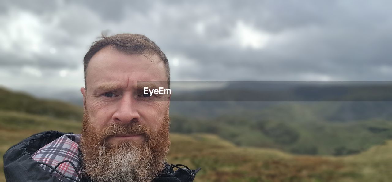one person, adult, portrait, beard, cloud, facial hair, men, sky, headshot, landscape, nature, mature adult, mountain, environment, overcast, looking at camera, person, scenics - nature, beauty in nature, emotion, front view, outdoors, lifestyles, focus on foreground, clothing, leisure activity, rural scene, activity, human face, day, human hair, contemplation, non-urban scene, serious, copy space, land, looking, travel, hiking, solitude