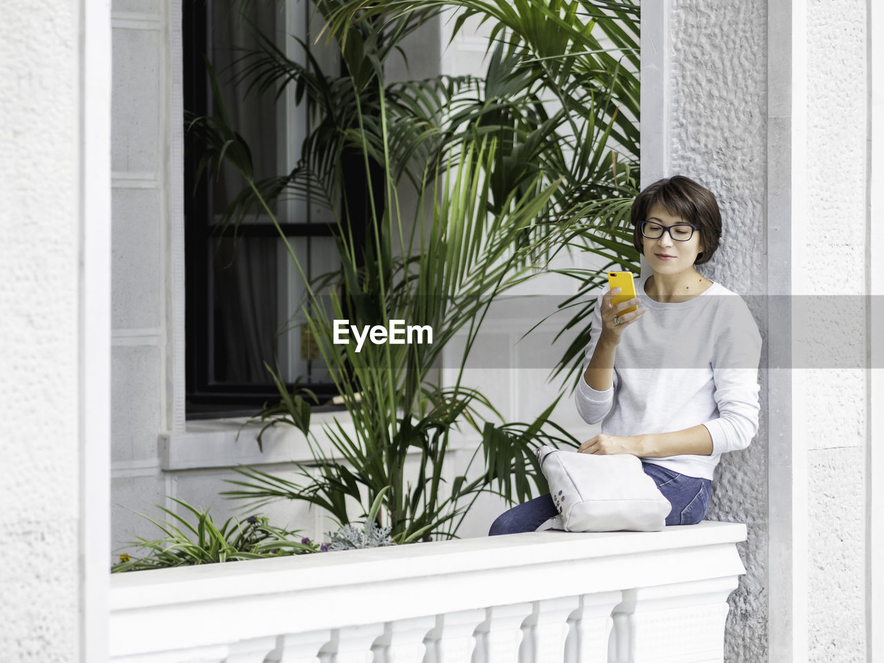 Woman texting on smartphone. pretty female in eyeglasses sits on balcony with palm trees. 