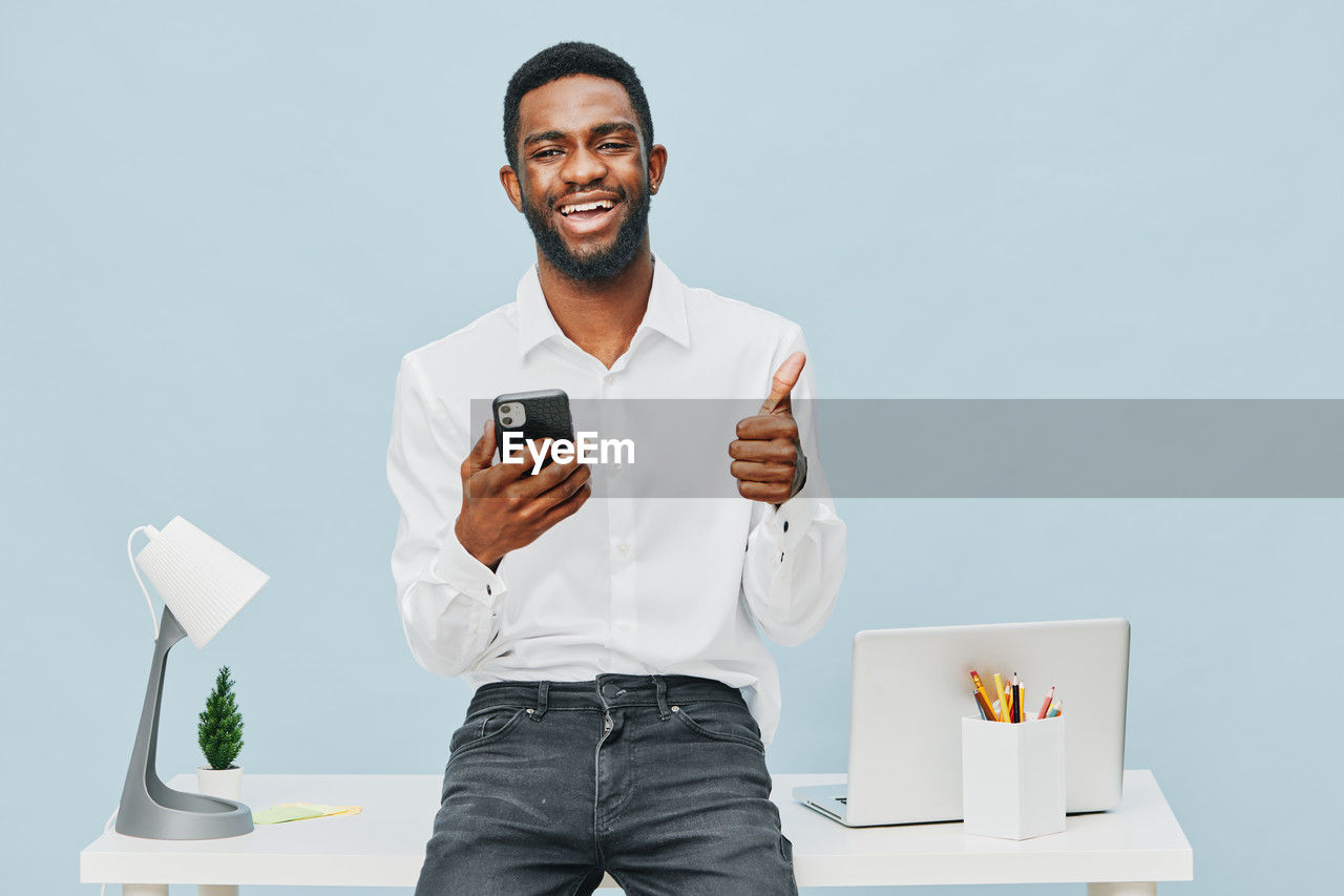one person, technology, adult, wireless technology, smiling, communication, men, happiness, smartphone, business, portable information device, young adult, mobile phone, indoors, front view, holding, emotion, portrait, studio shot, businessman, person, cheerful, copy space, telephone, business finance and industry, standing, computer, three quarter length, clothing, casual clothing, beard, internet, looking, computer network, portability, white-collar worker, occupation, facial hair, digital tablet, smile, gray, hand, teeth, looking at camera, table, new business, coffee, office, food and drink, lifestyles