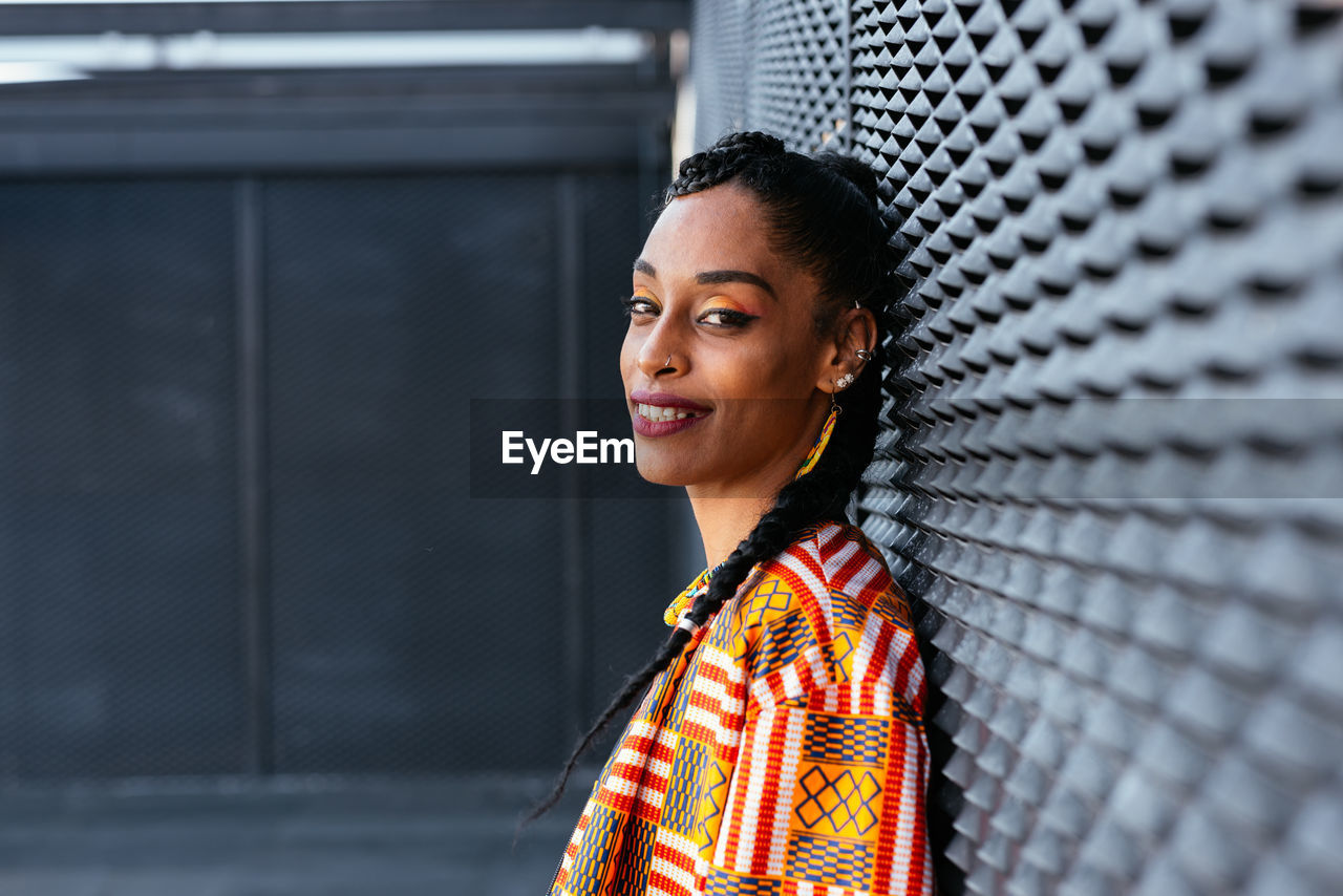 Side view of happy ethnic woman in colorful shirt with braid leaning on gray wall and looking at camera with smile in daytime on city street