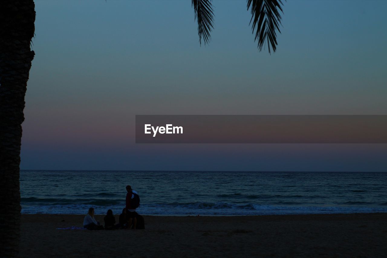 SILHOUETTE MEN SITTING ON BEACH AGAINST CLEAR SKY