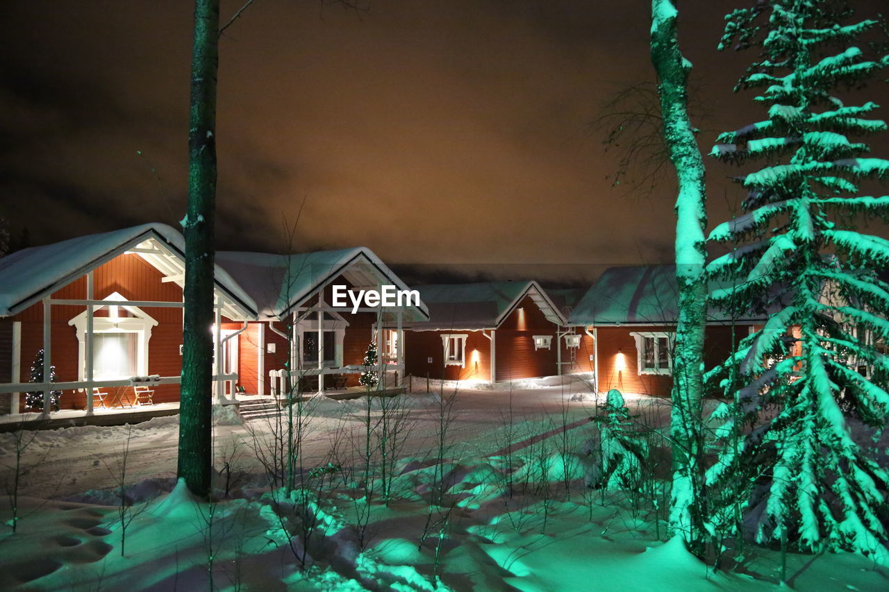 Snowy houses by bare trees against sky at night