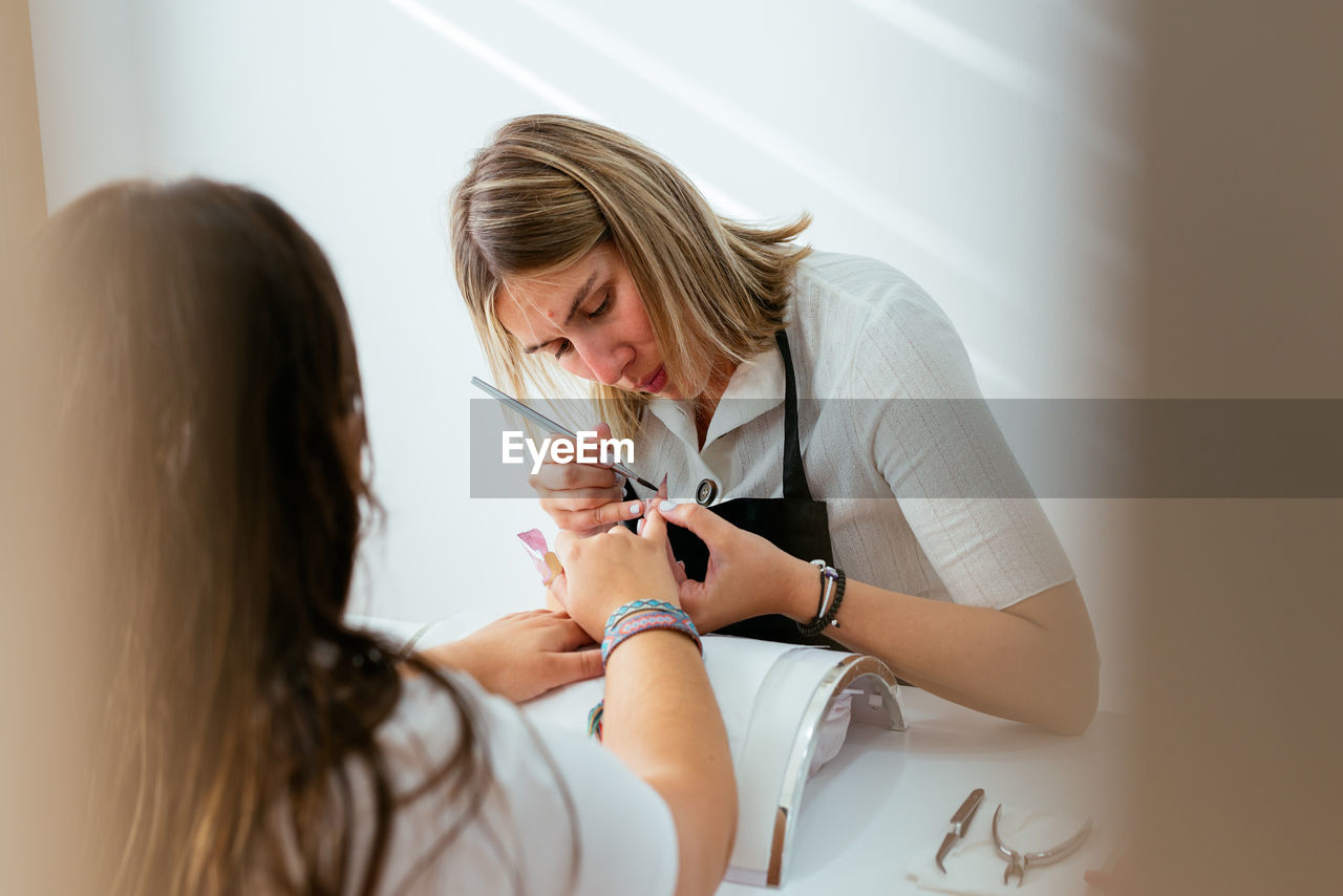 Concentrated female manicurist using brush while applying acrylic nail powder on tips of anonymous client in daylight in beauty salon