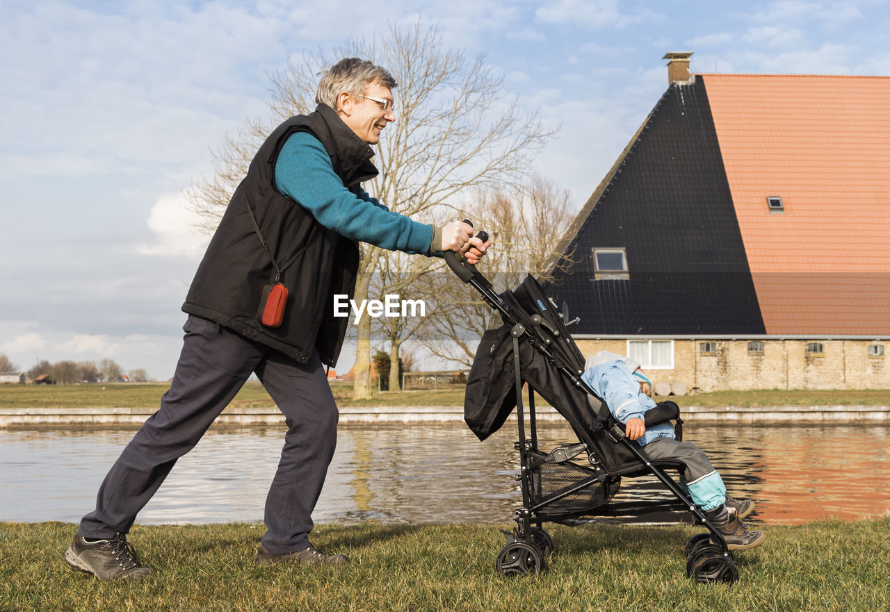Side view of grandfather pushing baby stroller on field by lake