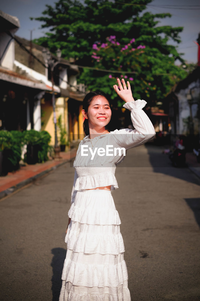 Smiling young woman waving while standing on footpath