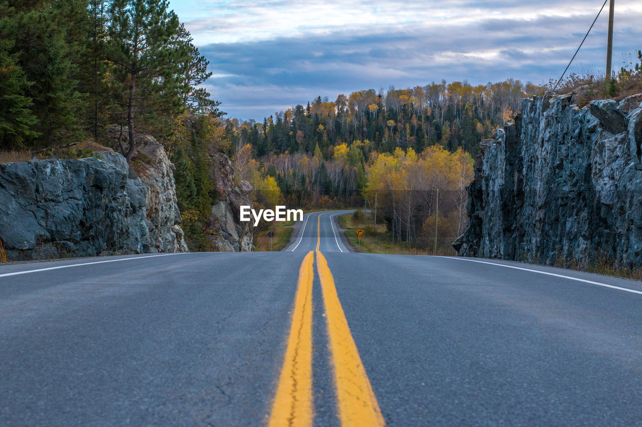 Empty road leading towards autumn trees against cloudy sky