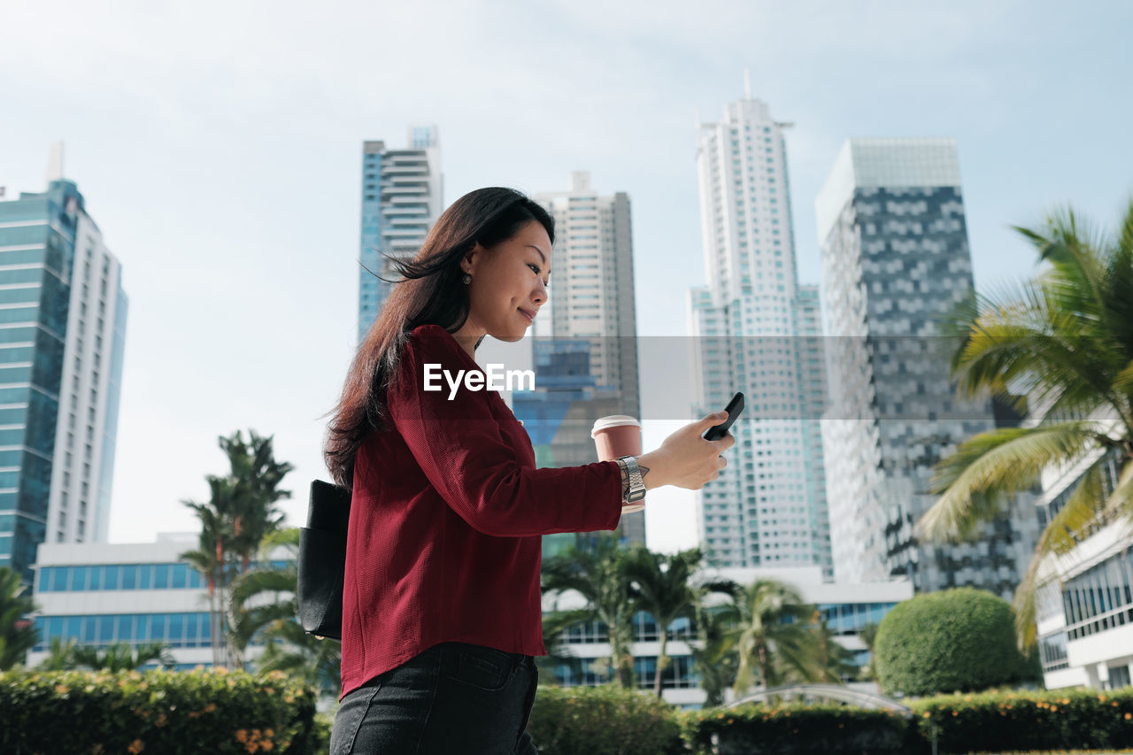 Side view of woman using smart phone while standing against buildings