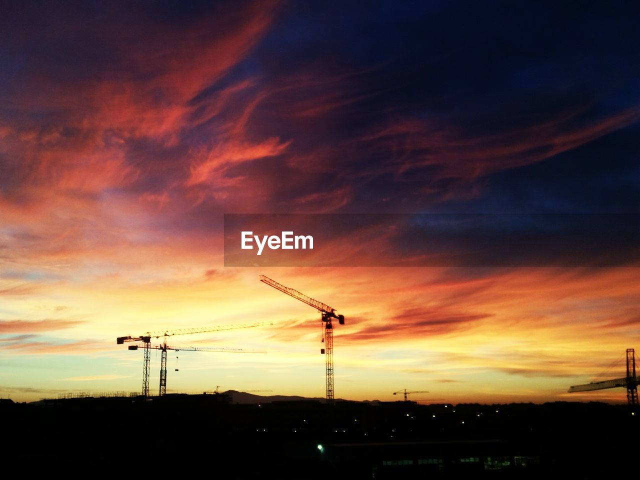 Silhouette cranes against cloudy sky during sunset