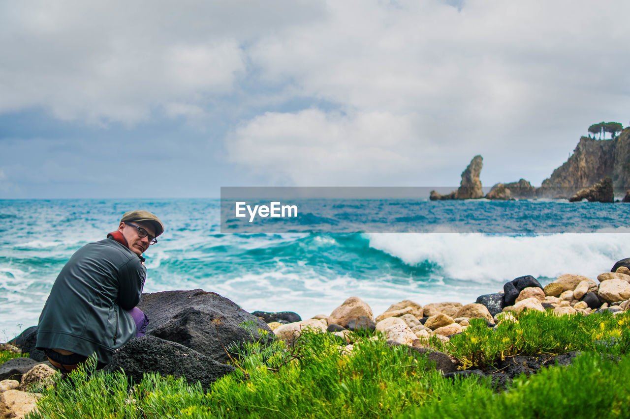 MAN SITTING ON ROCK AT SEA SHORE AGAINST SKY