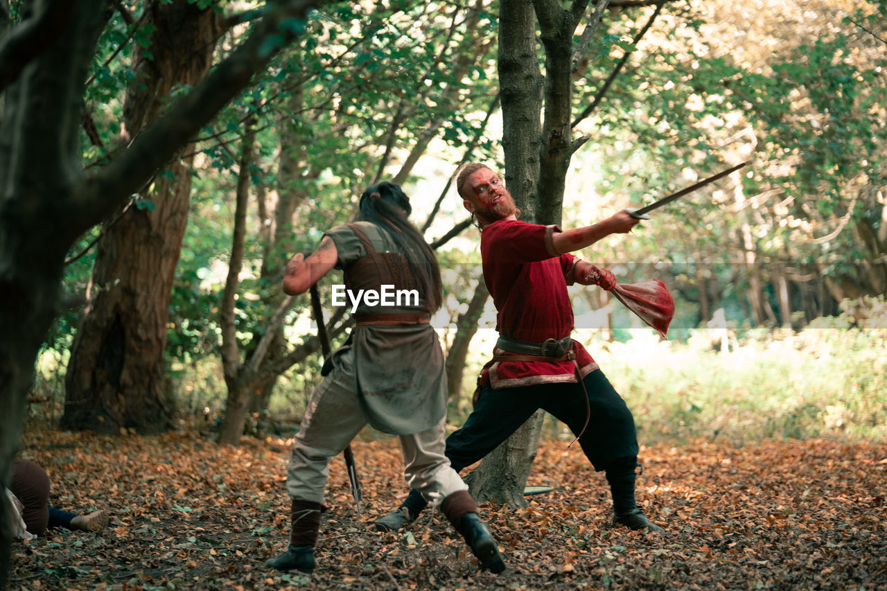 Full body of angry warrior in viking costume fighting opponent with sword during battle in forest
