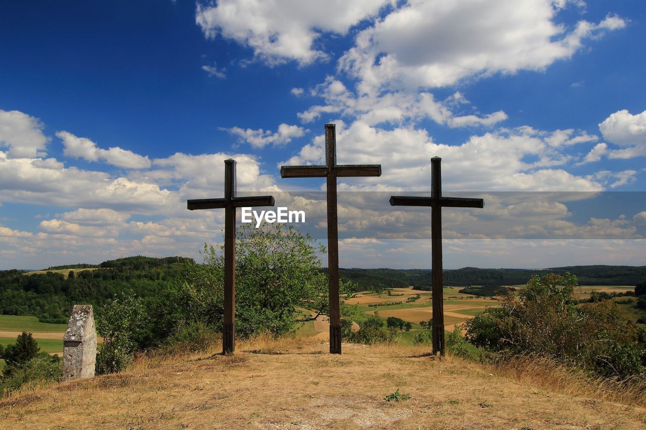 cross, religion, belief, spirituality, sky, cloud, catholicism, cross shape, nature, crucifix, grave, landscape, symbol, place of worship, environment, cemetery, tranquility, no people, land, idyllic, tranquil scene, beauty in nature, plant, outdoors, scenics - nature, sadness, hill, death, day, religious equipment, rural area, cloudscape, blue, grass, non-urban scene, architecture, stone, field, rural scene