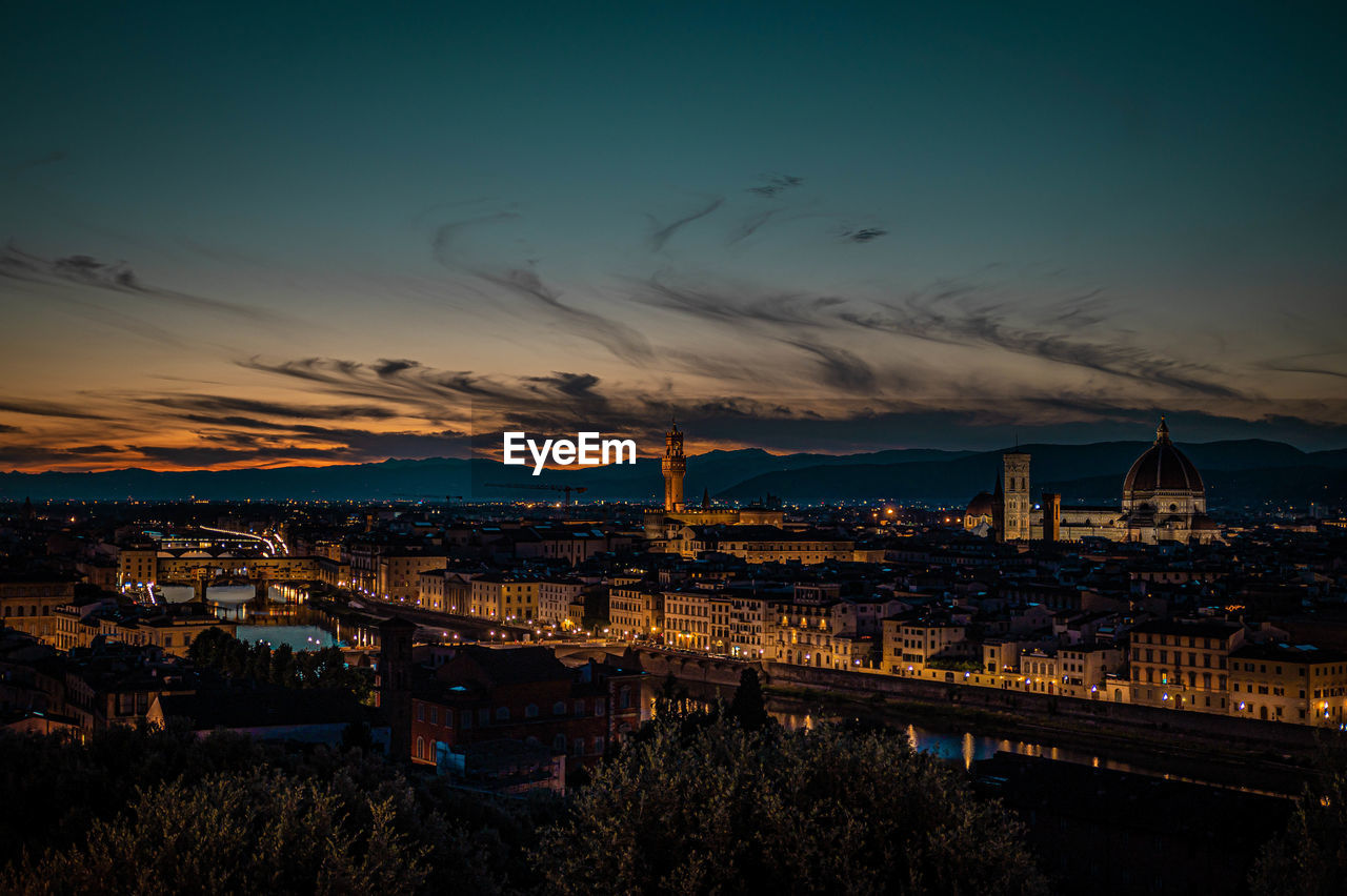 HIGH ANGLE VIEW OF ILLUMINATED BUILDINGS BY RIVER AGAINST SKY