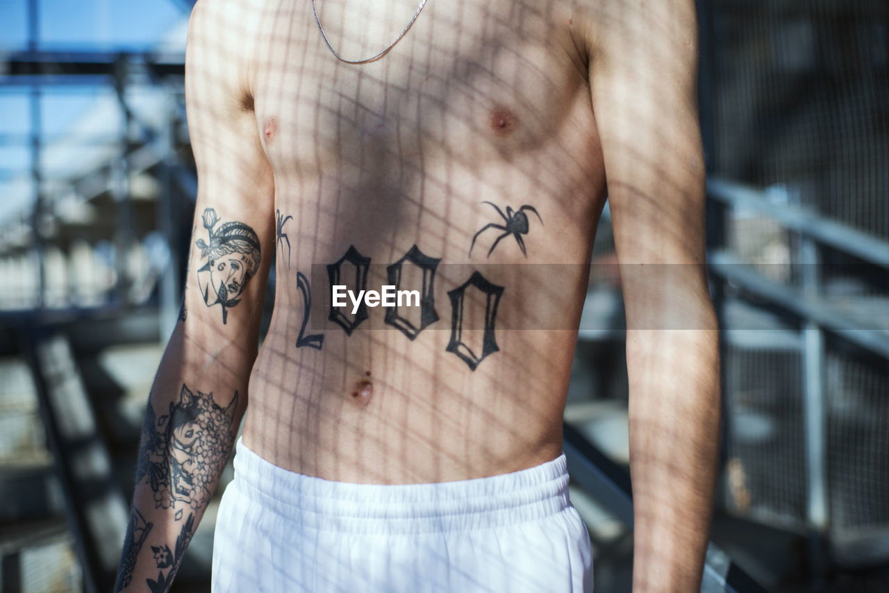 Midsection of shirtless man with tattoo in sunlight at home