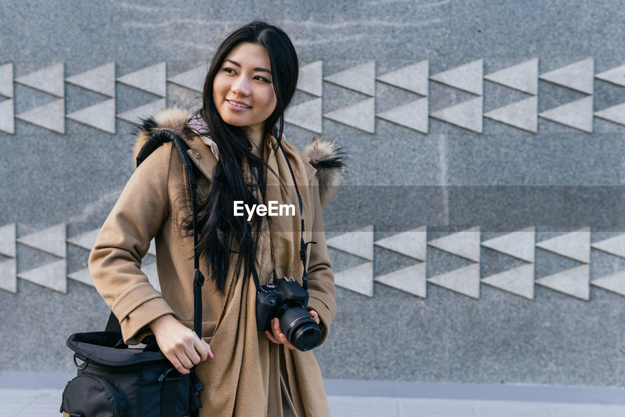 Positive asian female photographer wearing warm coat with photo camera standing against stone wall with geometric decor