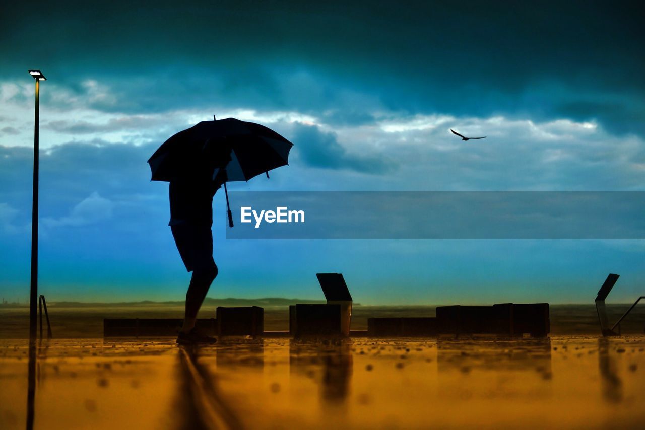 Silhouette man holding umbrella while walking at beach against cloudy sky during monsoon