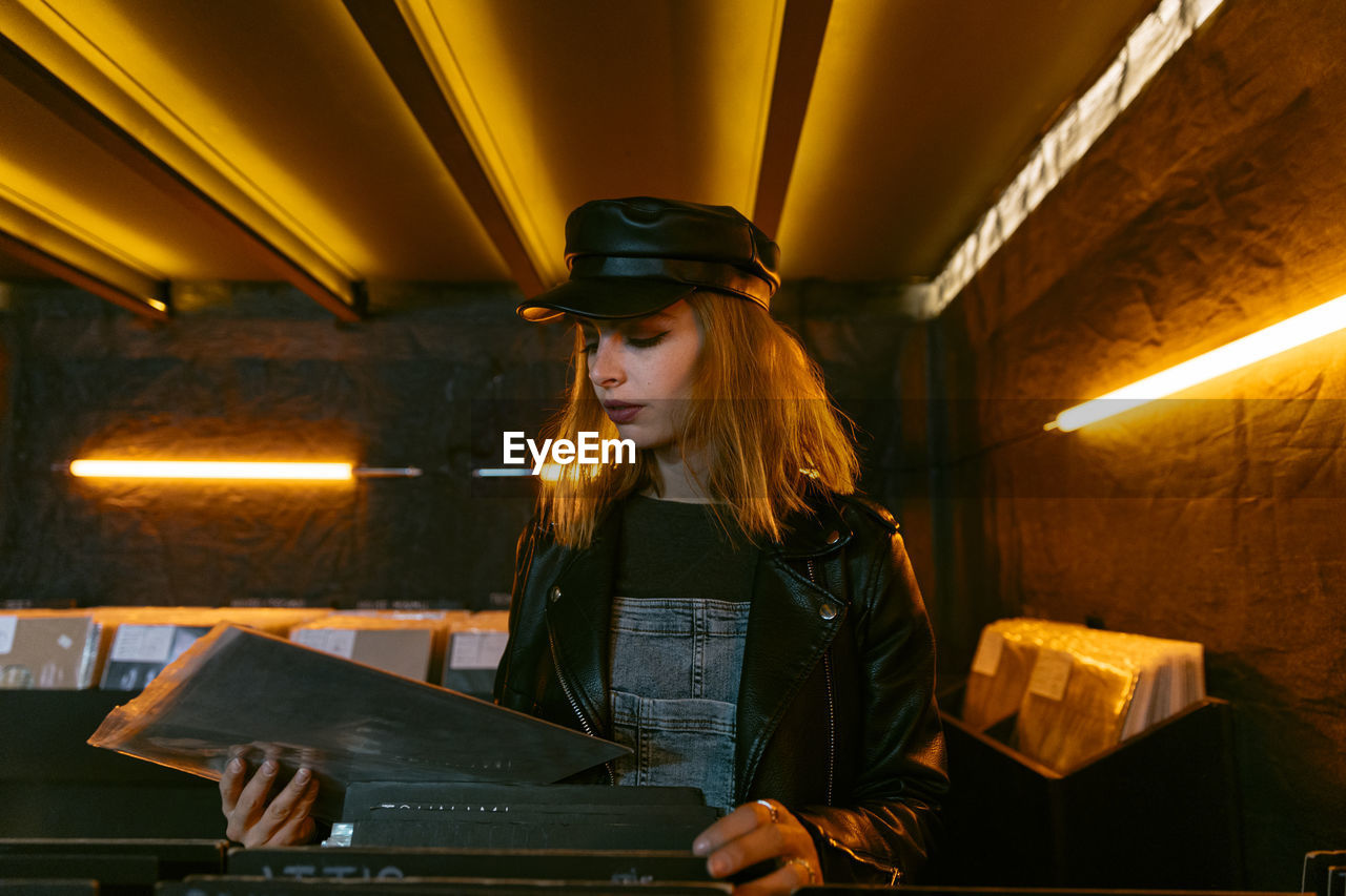 Tranquil young female in black cap choosing vinyl record while standing near counter with collection of musical albums in store with glowing lamps