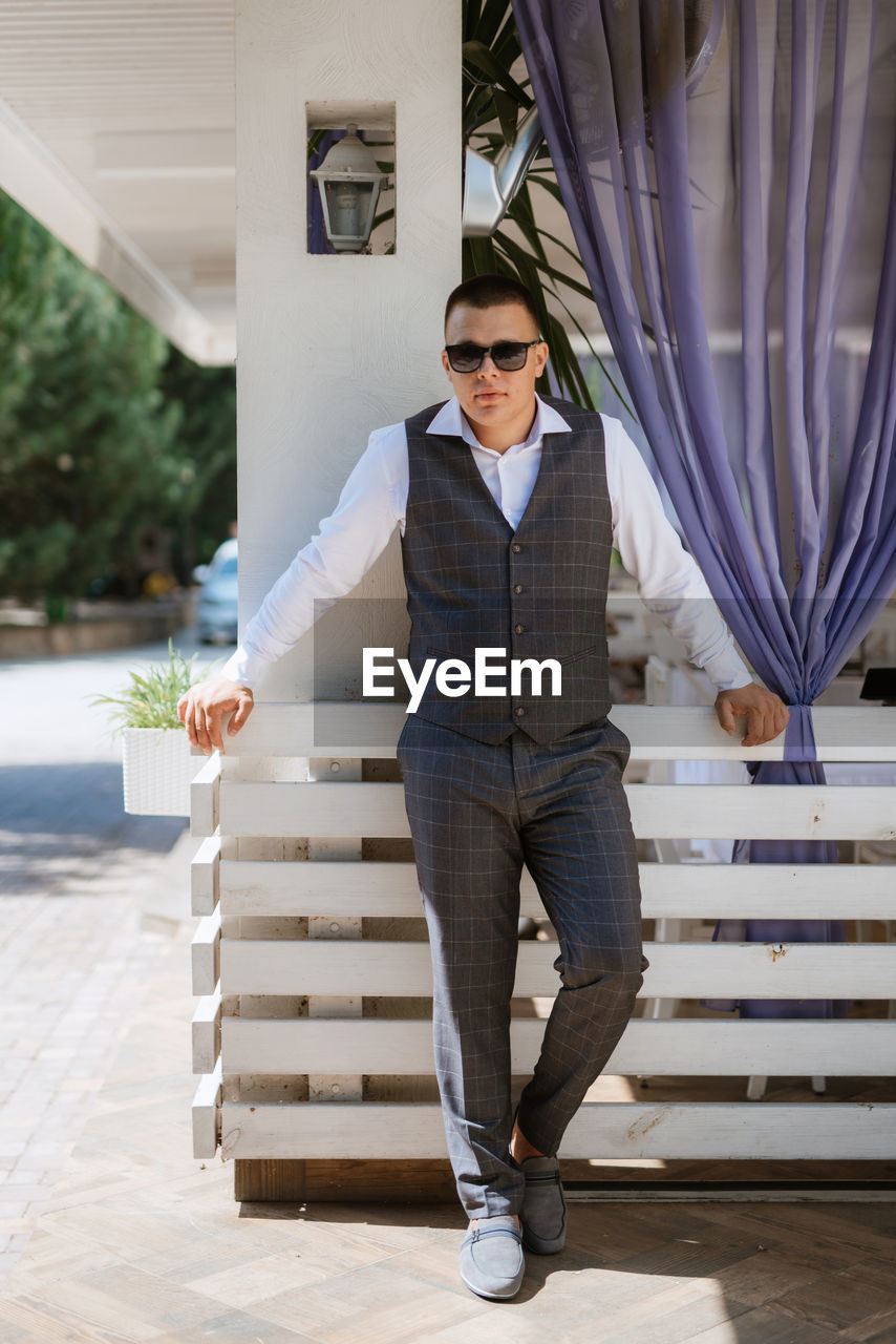 one person, adult, full length, men, glasses, fashion, sunglasses, architecture, front view, spring, standing, portrait, business, day, young adult, looking at camera, lifestyles, trousers, businessman, outdoors, staircase, formal wear, blue, holding, clothing, brown hair, casual clothing, smiling, built structure, emotion, sunlight