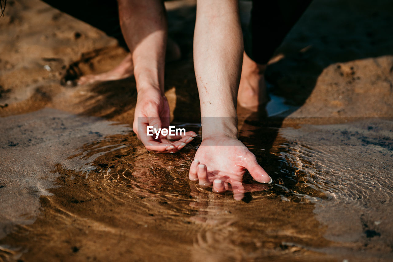 Unrecognizable tender female washing hands in crystal clear water of pond while sitting on sandy beach in summer