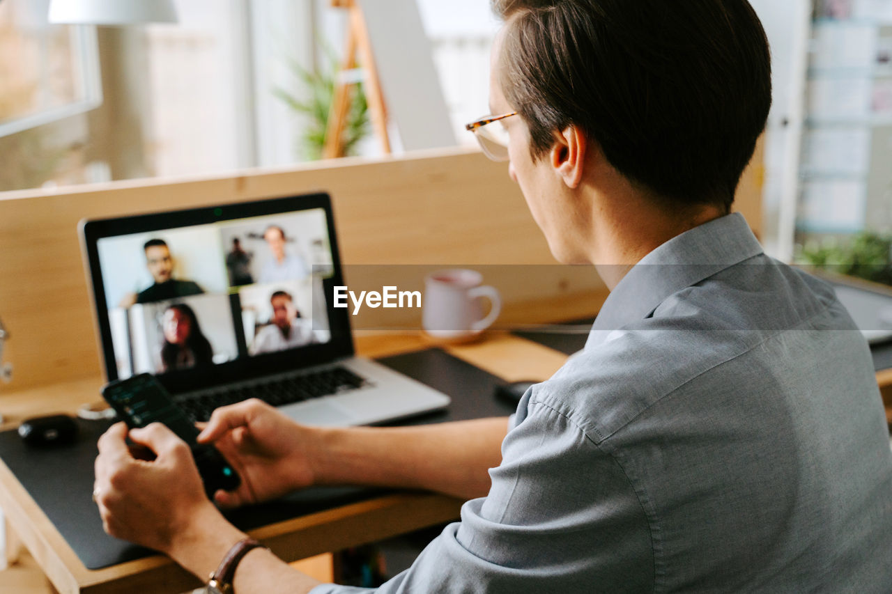 Side view of smiling male manager having video conference online via laptop while sitting at table and working from home during coronavirus epidemic