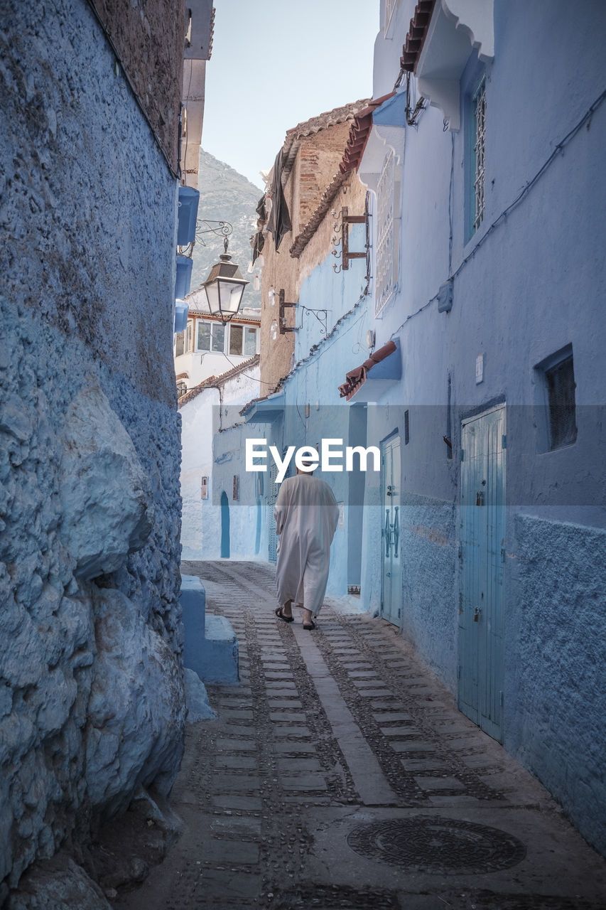 REAR VIEW OF WOMAN WALKING ON FOOTPATH BY BUILDINGS