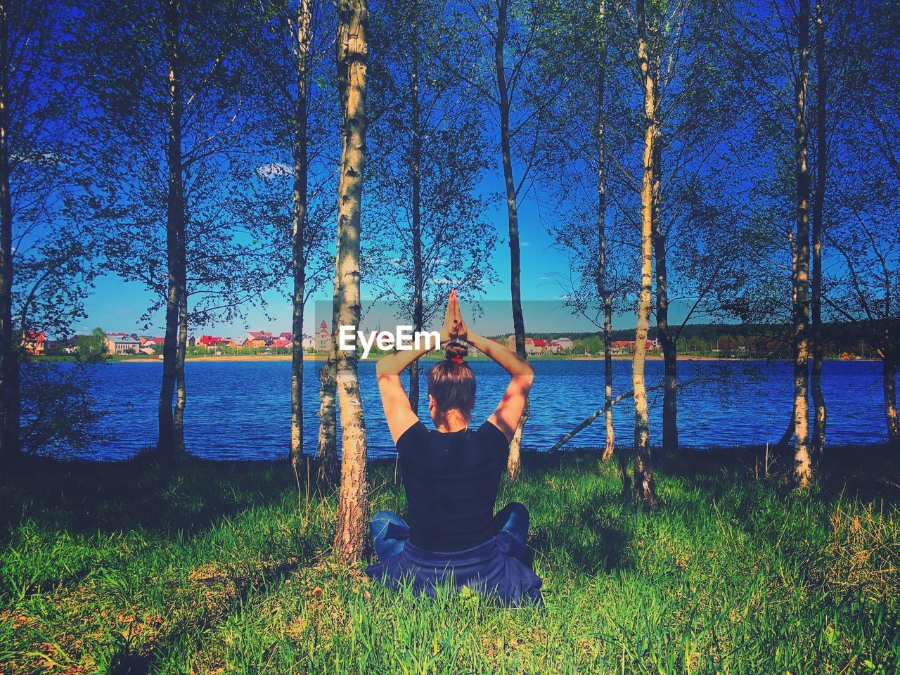 Rear view of woman meditating while sitting on grassy field by lake in forest