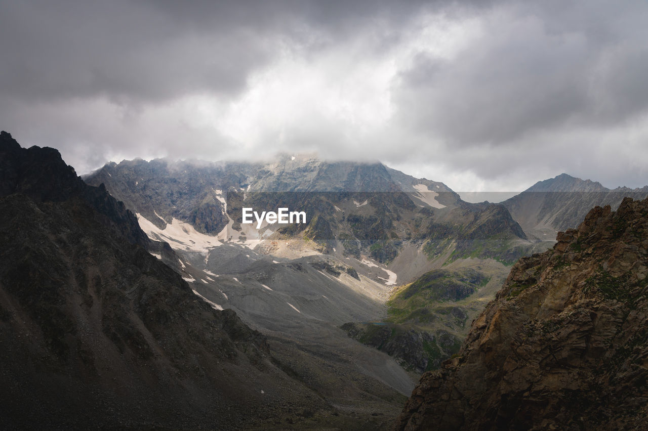 Dramatic view of forested high rocky slopes and snowy mountains in gray cloudy sky in changeable