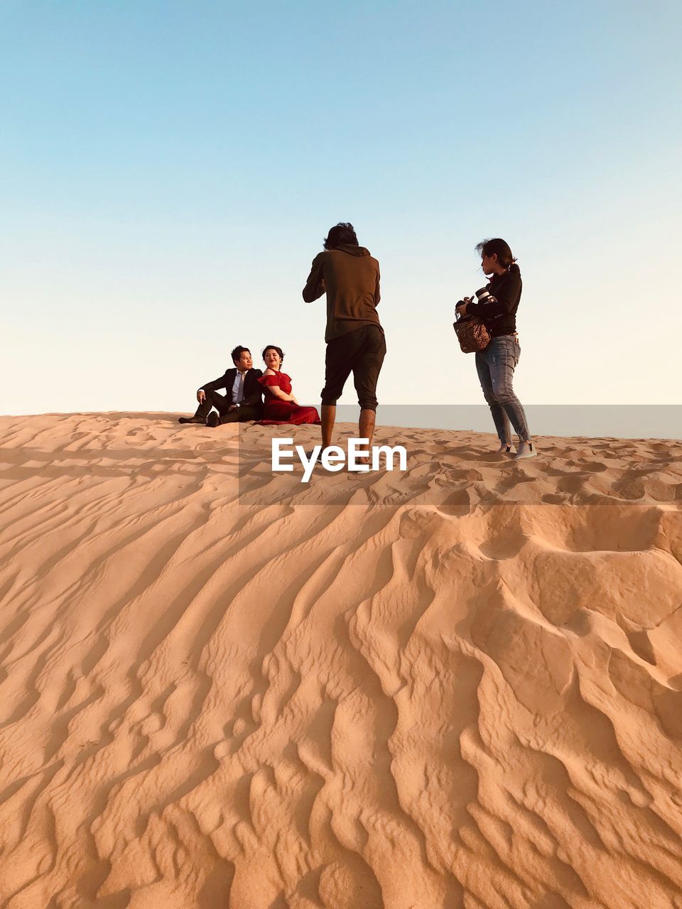 People photographing bride and groom sitting on sand against sky